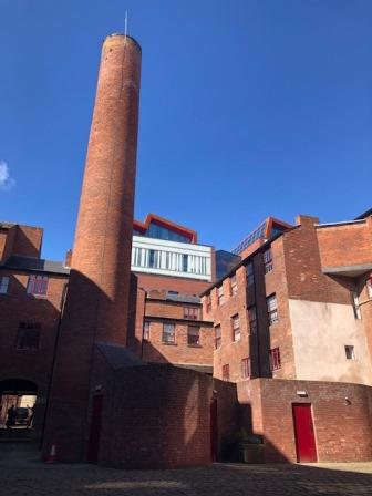The grade II listed Butcher Works in Sheffield on a sunny day with a clear blue sky. The top of  Sheffield Hallam University's Charles Building can be seen over the top of the old red brick buildings. 